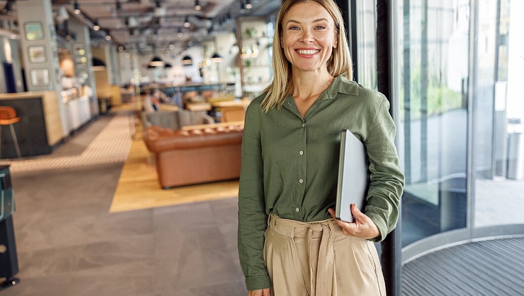 Eine Person steht in einem modernen Büro und hält einen Laptop in der Hand. Im Hintergrund sind Sofas und ein offener Arbeitsbereich zu sehen.